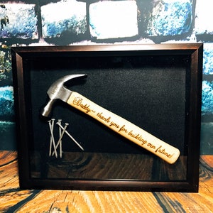 May include: A framed hammer with a wooden handle engraved with the text "Daddy - thank you for building our future!" and several nails are displayed on a black background.