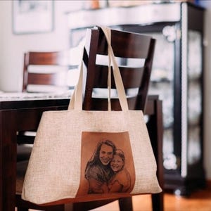 May include: A beige tote bag with long straps hangs on a wooden chair. The bag features a brown leather patch with a black and white portrait of two people. The bag is rectangular and appears to be made of a woven fabric.