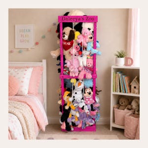 May include: A tall, bright pink wooden storage unit with the text "Daleeya's Zoo" engraved on the top frame. The unit features vertical white elastic cords creating a cage-like structure filled with a variety of colorful plush stuffed animals.