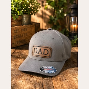 May include: A grey baseball cap featuring a rectangular tan leather patch on the front with the word DAD embossed in black capital letters. The cap has a curved brim with a circular sticker that reads The Original Flexfit XL/XXL.