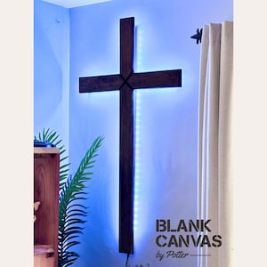 May include: A large, dark wooden cross mounted on a light blue wall, illuminated with blue LED lights. The cross is the focal point, with the text "BLANK CANVAS by Potter" in the lower right corner. A curtain and plant are visible.