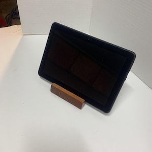 May include: A black tablet is propped up on a small, rectangular wooden stand. The tablet's screen is dark, reflecting the surrounding environment. The stand is a light brown color and appears to be made of wood.