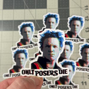 May include: A collection of stickers featuring a portrait of a man with blue hair. The stickers have the text "ONLY POSERS DIE" in a black, red, and white design. The stickers are on a cutting mat.