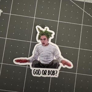 May include: A sticker featuring a cartoon drawing of a man with green hair, wearing a white shirt and red gloves. The text "GOD OR BOB?" is printed below the image.