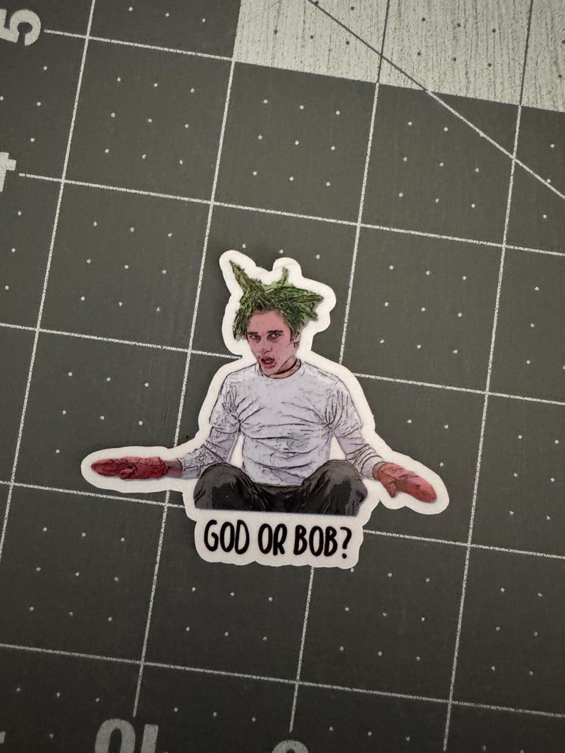 May include: A sticker featuring a cartoon drawing of a man with green hair, wearing a white shirt and red gloves. The text "GOD OR BOB?" is printed below the image.