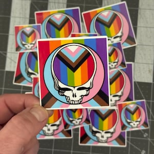 May include: A collection of square stickers featuring a white skull design within a rainbow-colored circle, set against a background of the Pride flag colors. The stickers are scattered on a gray surface.