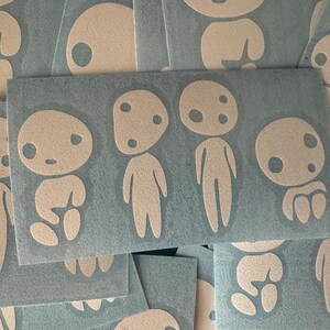 May include: Several white vinyl decal stickers featuring whimsical, minimalist character designs reminiscent of forest spirits. The kodama characters have simple, round bodies, small eyes, and no mouths.  They are arranged on a textured, dark-gray background. The stickers are ideal for adding a touch of whimsy to laptops, water bottles, or other smooth surfaces.