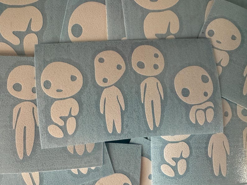 May include: Several white vinyl decal stickers featuring whimsical, minimalist character designs reminiscent of forest spirits. The kodama characters have simple, round bodies, small eyes, and no mouths.  They are arranged on a textured, dark-gray background. The stickers are ideal for adding a touch of whimsy to laptops, water bottles, or other smooth surfaces.