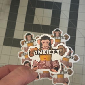 May include: A collection of stickers featuring a cartoon monkey with the word "ANXIETY" printed above it. The monkey has a brown face, yellow torso, and red and white striped pants. The monkey is holding a brown object in each hand.