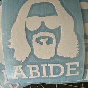 May include: A blue sticker with a white silhouette of a man with long hair and a beard wearing sunglasses. The text "ABIDE" is printed below the silhouette.