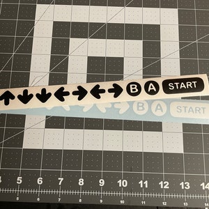 May include: A set of black and white stickers for a video game controller. The stickers include directional arrows, buttons labeled "A", "B", and "Start".