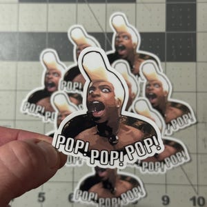 May include: A collection of stickers featuring a person with a surprised expression and a tall, white hairstyle. The stickers have the word "POP!" repeated multiple times. The stickers are held by a hand.