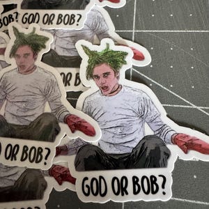 May include: A set of stickers featuring a cartoon character with green hair and the text "God or Bob?" The character is wearing a white shirt and red mittens.