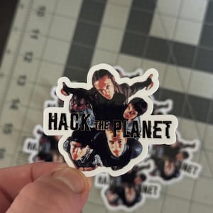 May include: A sticker featuring a group of people with the text "HACK THE PLANET" in a bold, black font. The sticker has a white border and is likely made of vinyl.