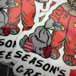 May include: Two red and white Christmas stickers featuring a character in a red jumpsuit holding a sack and a stick. The stickers have the text "Season's Greetings" on them.