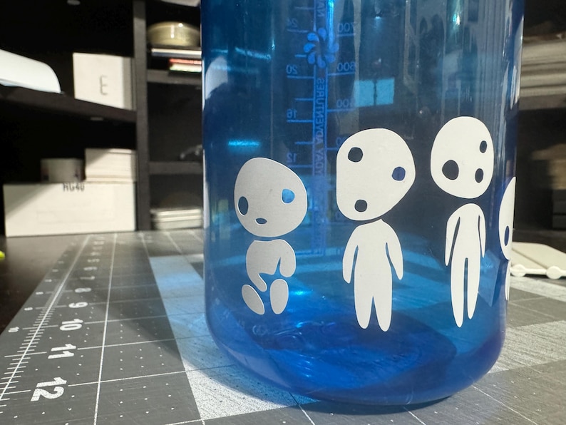 May include: Blue water bottle with white cartoon characters on the side. The characters have large eyes and round bodies. The bottle has a measurement scale on the side.