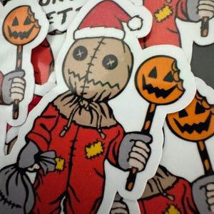 May include: A red and brown Halloween sticker of a character wearing a Santa hat and holding a candy cane with a bitten pumpkin on the end. The character has a stitched face and is wearing a red jumpsuit with yellow patches.