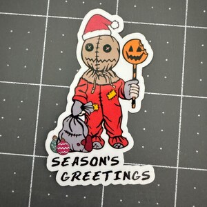 May include: A red and white Christmas-themed sticker featuring a stitched-faced character wearing a Santa hat and holding a candy cane with a bitten pumpkin on top. The sticker says "Season's Greetings".