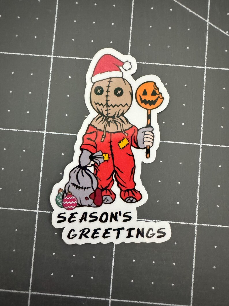 May include: A red and white Christmas-themed sticker featuring a stitched-faced character wearing a Santa hat and holding a candy cane with a bitten pumpkin on top. The sticker says "Season's Greetings".