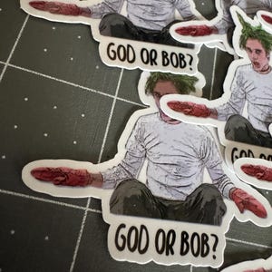 May include: Four stickers featuring a cartoon character with green hair and a white shirt. The character is holding a red object in their hand. The stickers have the text "God or Bob?" printed on them.