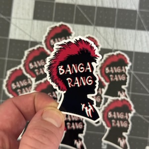 May include: A black sticker featuring a silhouette of a person with red hair and the text "BANGA RANG" in white. The sticker has a 3D effect with red and blue accents. Additional stickers are visible in the background.