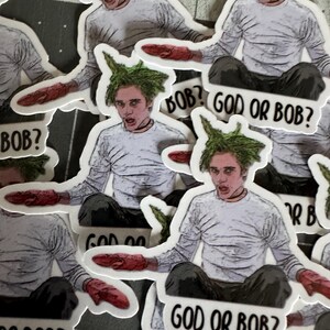 May include: A set of stickers featuring a cartoon character with green hair and a white shirt. The character is holding a red object in their hand. The stickers have the text "God or Bob?" printed on them.