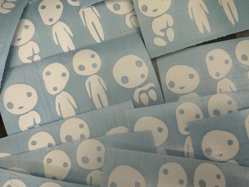 May include: A close-up of a pile of blue vinyl stickers featuring white outlines of a character with two eyes and a mouth. The character is in various poses and sizes.