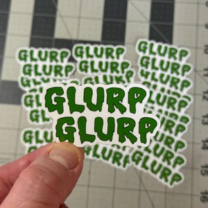 May include: A collection of white stickers with the word "GLURP" in a green, dripping font. The stickers are on a cutting mat with a grid pattern. The word "GLURP" is repeated multiple times.