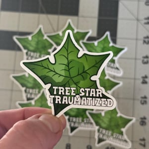 May include: A collection of stickers featuring a green leaf design with the text "TREE STAR TRAUMATIZED" in white. The stickers are held in a hand, with several others visible in the background. The stickers are on a cutting mat.