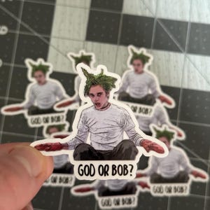 May include: A sticker featuring a cartoon character with green hair and a white shirt. The character is holding a red object in each hand. The sticker has the text "God or Bob?" printed on it.