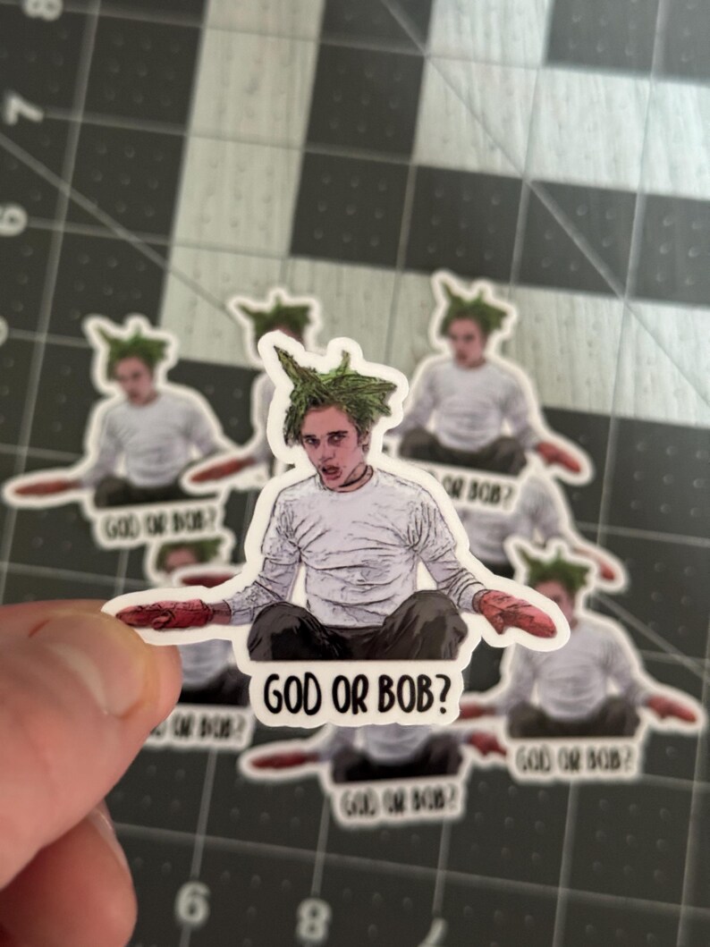 May include: A sticker featuring a cartoon character with green hair and a white shirt. The character is holding a red object in each hand. The sticker has the text "God or Bob?" printed on it.