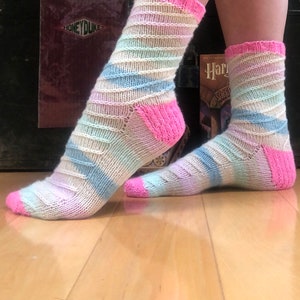 Può includere: Un paio di calzini a righe pastello con talloni e punte rosa. I calzini sono realizzati in un morbido tessuto a maglia e presentano una combinazione di colori blu chiaro, rosa e bianco.