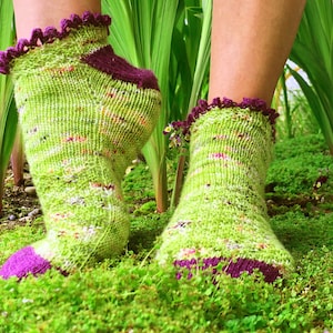 May include: A pair of green and purple knitted socks with a lacy trim. The socks are worn on a person's feet and are resting on a bed of green moss.