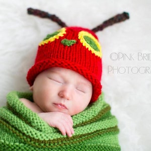 A baby boy with a red caterpillar hat on his head. Brown antennae point in both directions. his right hand is sticking out of a green rumpled knit swaddle sack while he sleeps. On a white background.