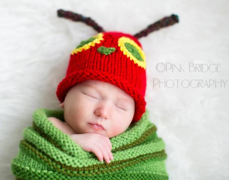 A baby boy with a red caterpillar hat on his head. Brown antennae point in both directions. his right hand is sticking out of a green rumpled knit swaddle sack while he sleeps. On a white background.