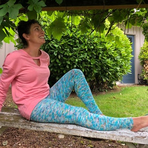 A woman sitting on a bench smiling up at the grape canopy above her. She is bear-footed, wearing a blue variegated knit legging and pink top. The leggings are garter stitch knit with ribbed ankles.