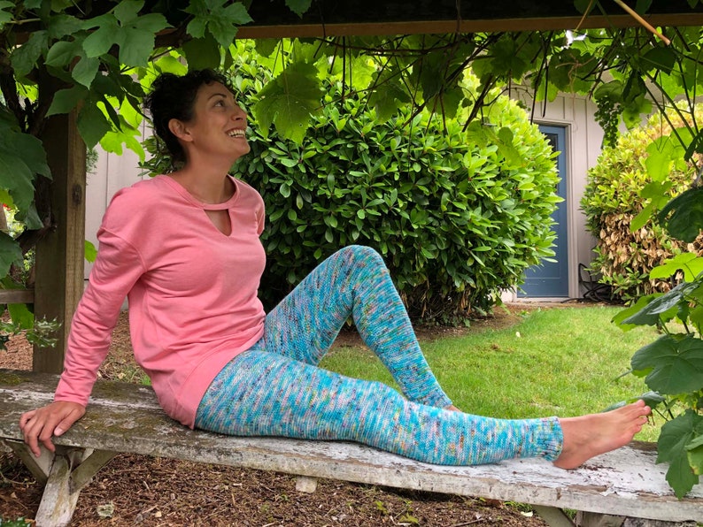 A woman sitting on a bench smiling up at the grape canopy above her. She is bear-footed, wearing a blue variegated knit legging and pink top. The leggings are garter stitch knit with ribbed ankles.