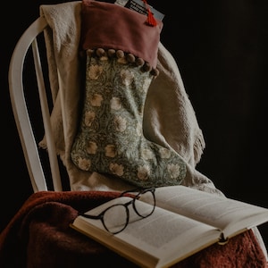 Op de afbeelding: Een kerstsok met bloemenpatroon, een bordeauxrode manchet en pompom-afwerking hangt aan een witte stoel. Een boek met een bril en een kaart met rode kwast liggen ook op de stoel, wat een gezellige scène creëert.