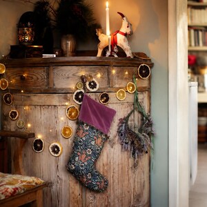 May include: A rustic wooden cabinet decorated for the holidays. A lit candle and a decorative goat figurine sit on top. A Christmas stocking with a floral pattern hangs on the front, along with a garland of dried citrus slices and fairy lights.