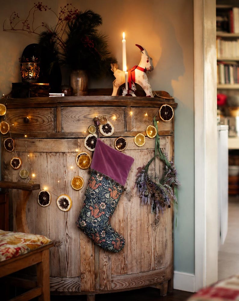 May include: A rustic wooden cabinet decorated for the holidays. A lit candle and a decorative goat figurine sit on top. A Christmas stocking with a floral pattern hangs on the front, along with a garland of dried citrus slices and fairy lights.
