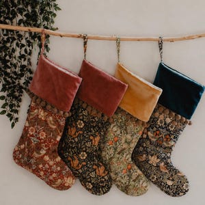 Puede incluir: Cuatro calcetines navideños cuelgan de una rama de madera. Cada calcetín tiene un estampado floral diferente y un puño de terciopelo en tonos rojos, dorados y verde azulado. Decorados con pompones.