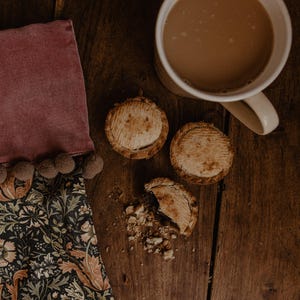 May include: Overhead shot of a wooden table with a mug of tea, three small pies, and a decorative cloth. The pies have a golden-brown crust. The cloth is dark with a floral pattern and a pink border with pom-poms.