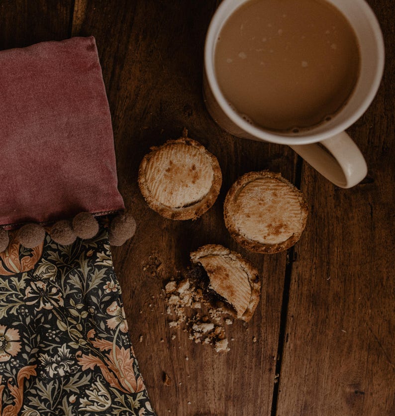 May include: Overhead shot of a wooden table with a mug of tea, three small pies, and a decorative cloth. The pies have a golden-brown crust. The cloth is dark with a floral pattern and a pink border with pom-poms.