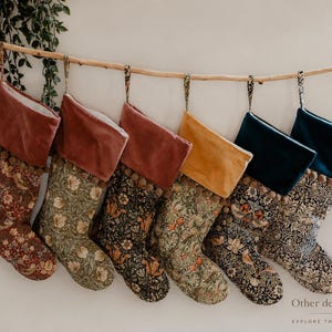 May include: Six Christmas stockings hanging from a wooden branch. The stockings have floral patterns in various colors, with velvet cuffs in shades of red, yellow, and teal. The text "Other designs available" is visible.