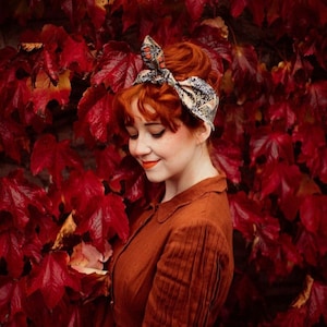 May include: A woman with red hair and a patterned headband smiles in front of a backdrop of red leaves. She wears a rust-colored dress with vertical pleats on the sleeves. The headband features a mix of colors and patterns.