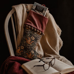 Puede incluir: Un calcetín navideño con estampado floral y puño de terciopelo, adornado con pompones. El calcetín está lleno de un libro, descansando sobre una silla con una manta. Un par de gafas y un libro abierto están en primer plano.