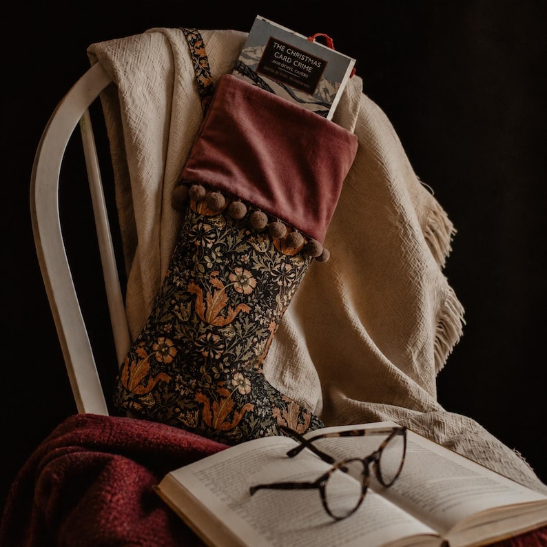May include: A Christmas stocking with a floral pattern and a velvet cuff, adorned with pom-poms. The stocking is filled with a book, resting on a chair with a blanket. A pair of glasses and an open book are in the foreground.