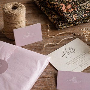May include: A still-life shot of packaging materials and stationery on a wooden surface. Includes a roll of twine, pink tissue paper, business cards, and a card with the word "Hello". A folded fabric with a floral pattern is also present.