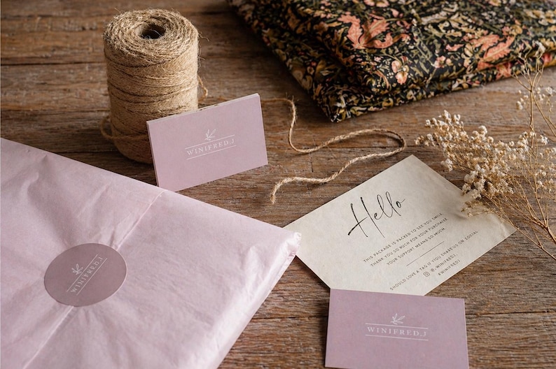 May include: A still-life shot of packaging materials and stationery on a wooden surface. Includes a roll of twine, pink tissue paper, business cards, and a card with the word "Hello". A folded fabric with a floral pattern is also present.