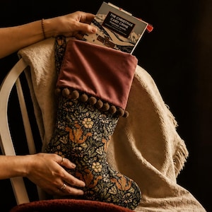 May include: A Christmas stocking with a floral and squirrel pattern, topped with a mauve velvet cuff and brown pom-poms. The stocking is filled with a book titled "The Christmas Card Crime". The stocking is resting on a chair.